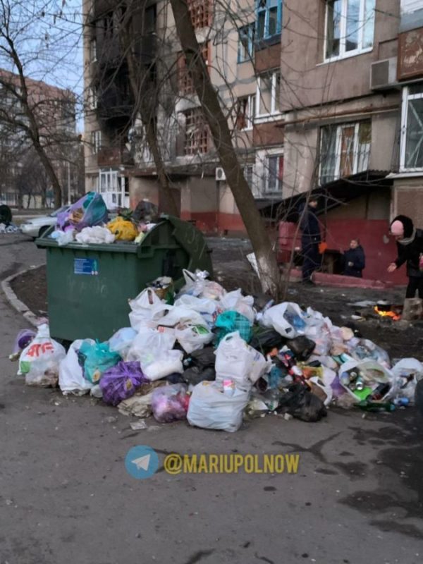 A woman cooking food on a bonfire not far from an overflown dumpster as people stand at the exit from the basement. Mariupol, Orlyka St. Published on 20 March 2022. Source. ~