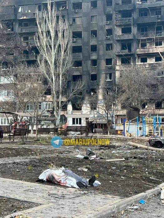 Dead body lying in front of charred buildings on Pylypa Orlyka St in Mariupol. Source. ~