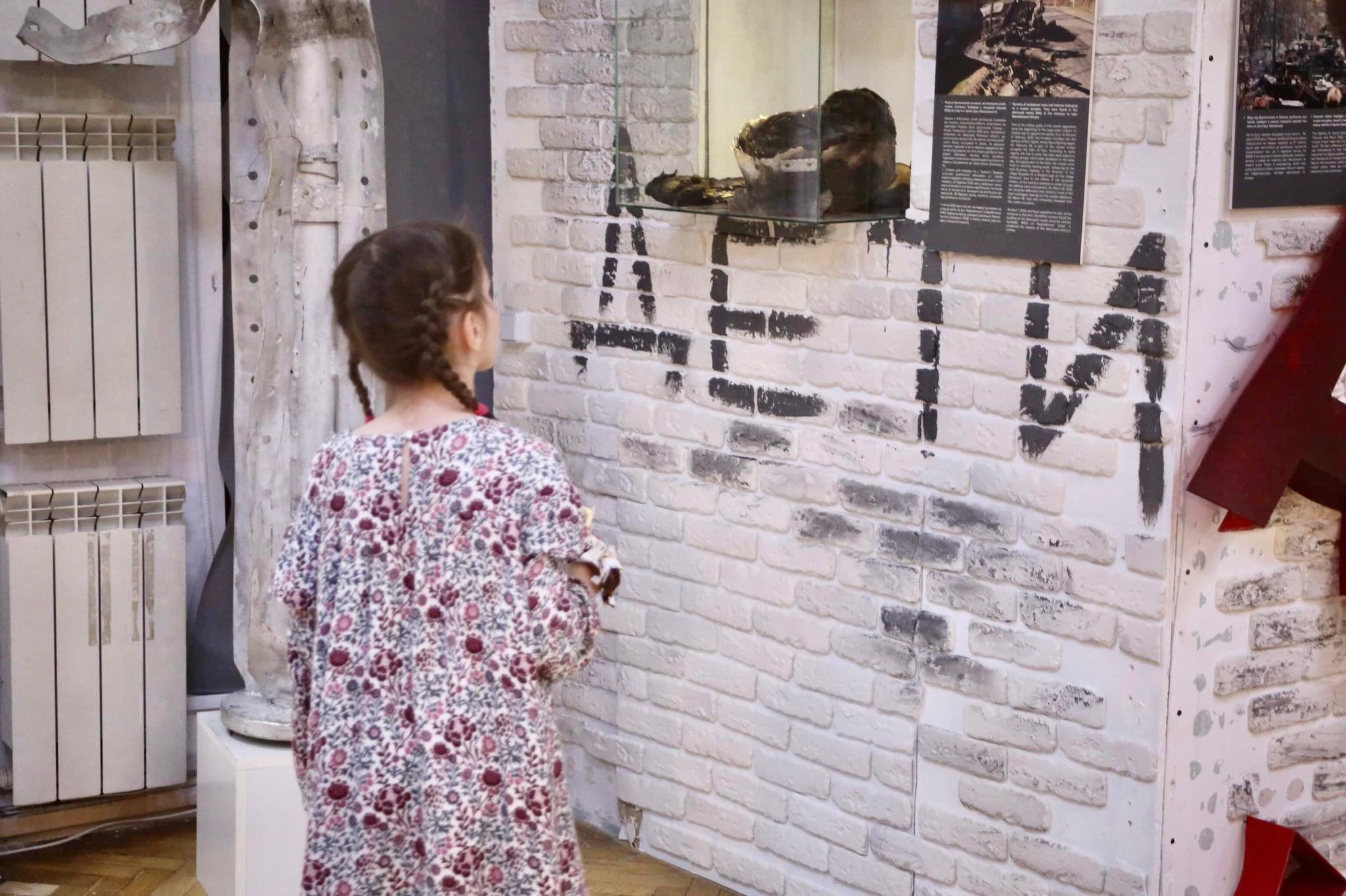 A little girl visits the exhibition “Invasion. Kyiv shot,” dedicated to the 2022 defense of Kyiv from the Russian invaders. The inscription on the wall – “Children” – was often written by the locals in places under Russian attack in a hope that a place is not hit. Photo by Euromaidan Press ~